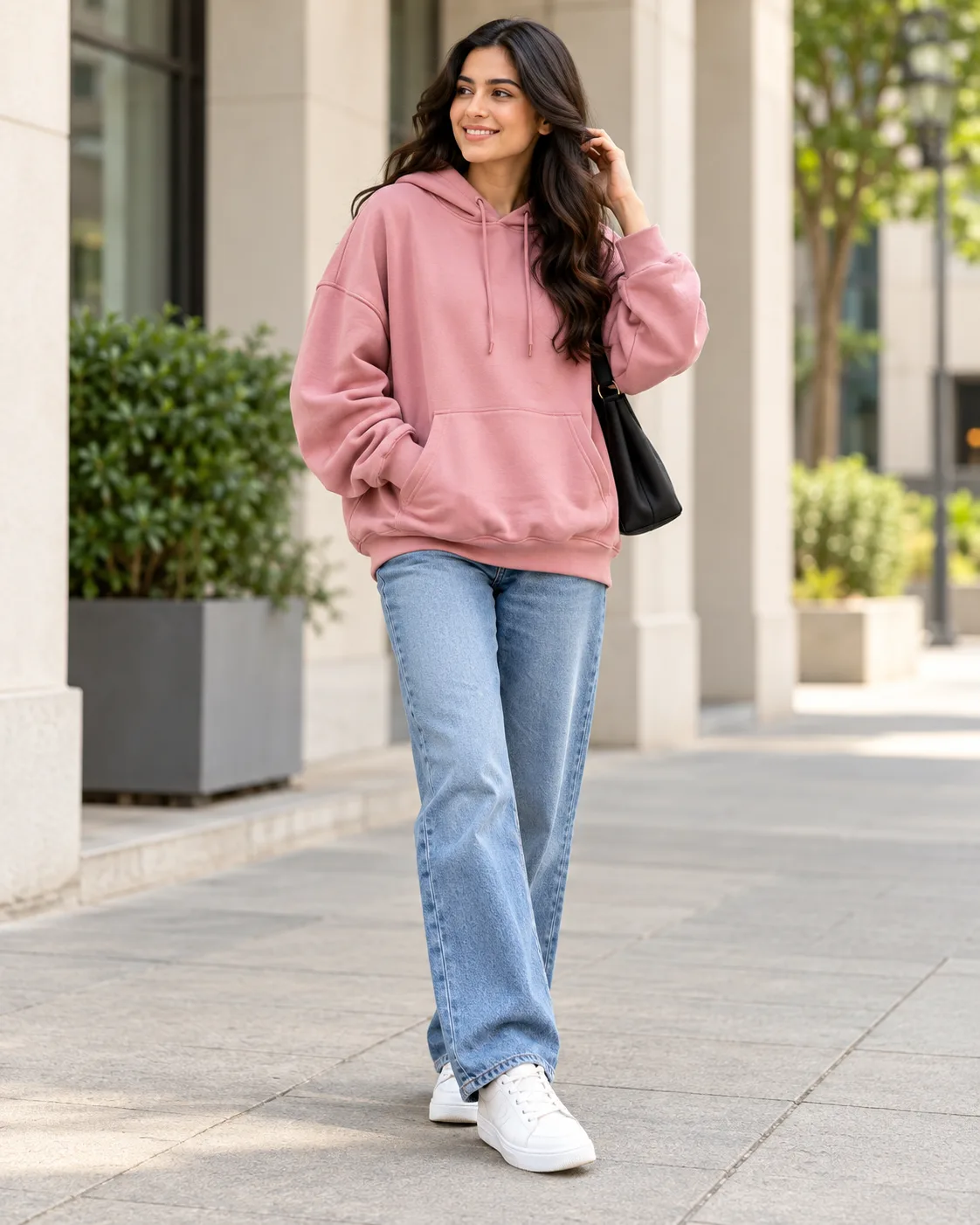 Woman wearing a pink oversized hoodie, straight-leg blue jeans and white sneakers on an urban walkway